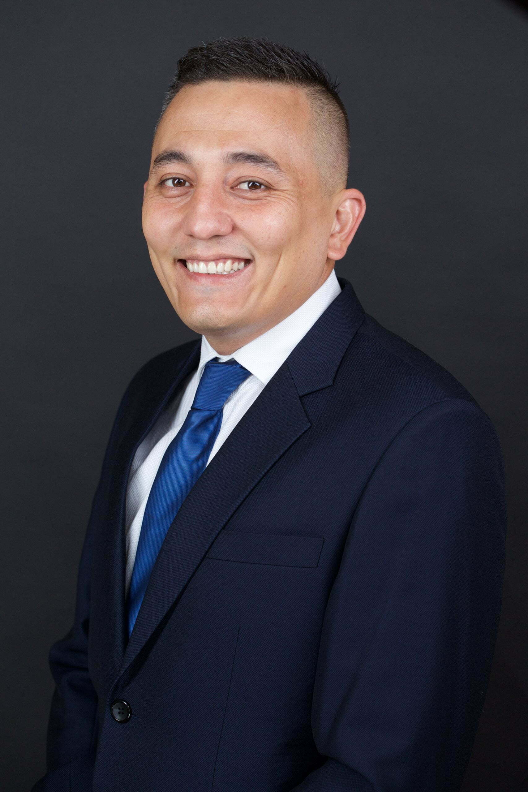 A man in a suit and blue tie smiling against a dark background.