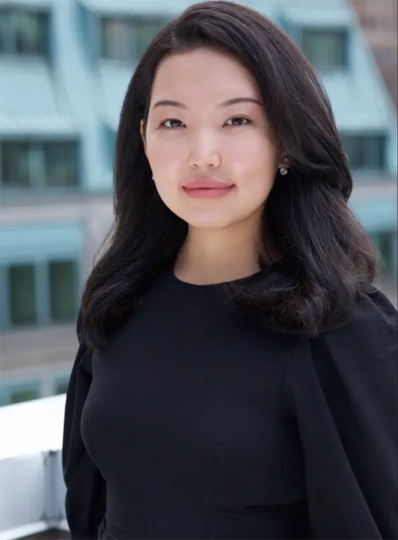 A woman with long black hair wearing a black top, standing outdoors with a blurred building in the background.