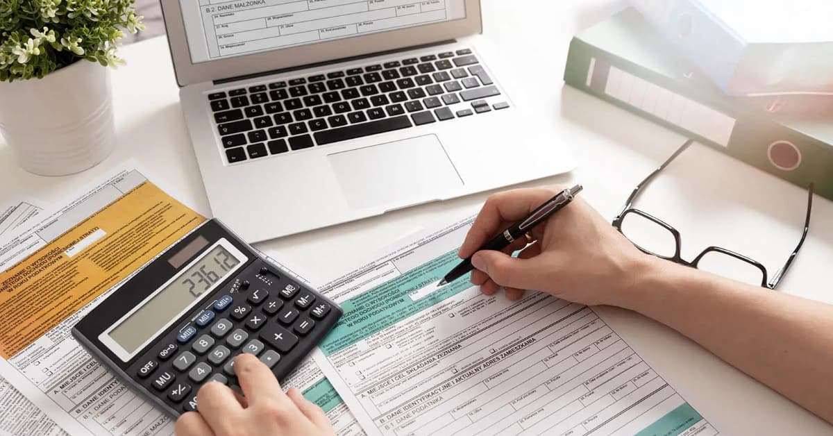 Person working on tax documents with a laptop, calculator, pen, and glasses on a desk.