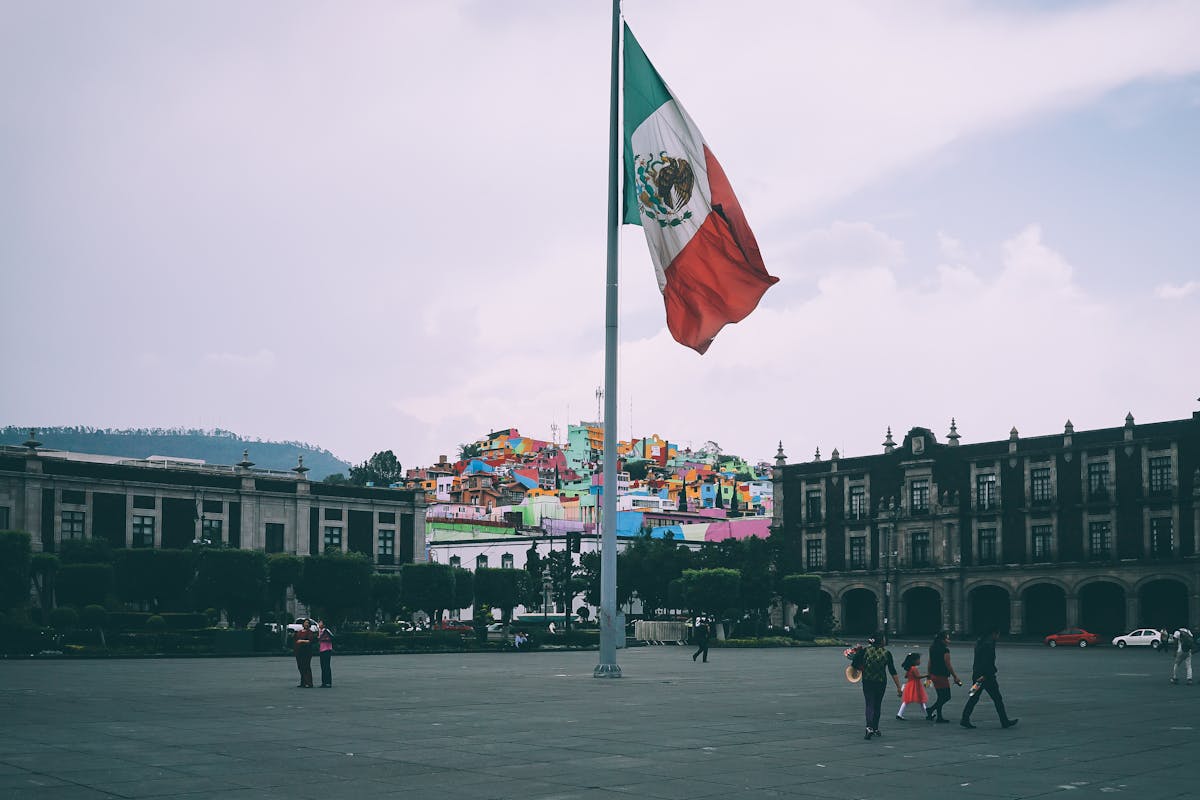 Large Mexican flag flying over a public square with historic buildings, representing residency, rights, and dual citizenship in Mexico.